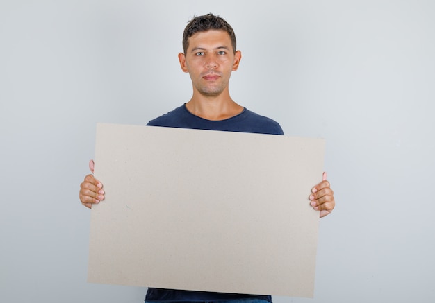 Young man holding blank poster in dark blue t-shirt and looking hopeful. front view.