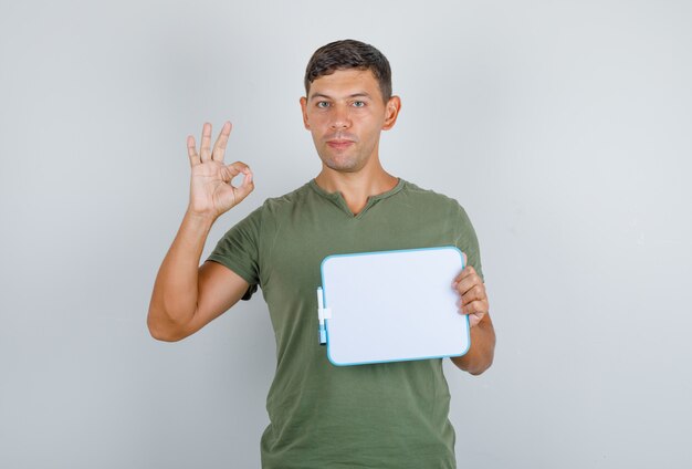 Young man holding board and doing ok sign in army green t-shirt front view.