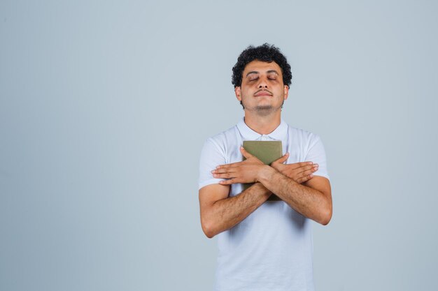 Young man holding book in white t-shirt and looking peaceful. front view.