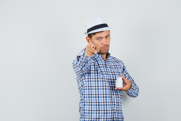 Young man holding bottle of pills, pointing at camera in checked shirt