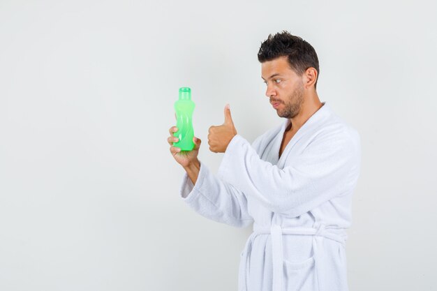 Young man holding bottle of water with thumb up in white bathrobe front view.