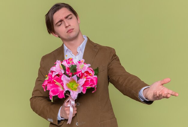 緑の壁の上に立っている国際女性の日を祝うために腕を上げて混乱して脇に花の花束を持っている若い男