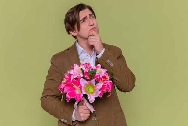緑の壁の上に立っている国際女性の日の概念を祝福するつもりである物思いにふける表現で見上げる花の花束を持っている若い男