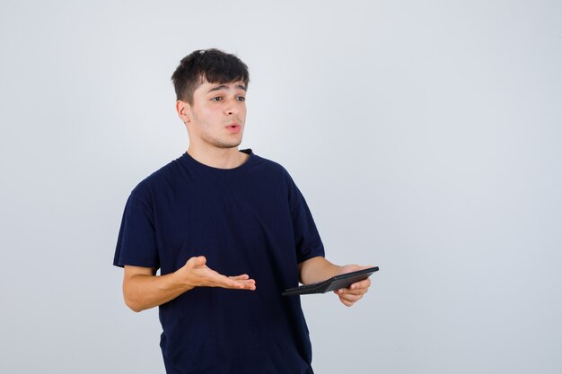 Young man holding calculator in black t-shirt and looking bewildered , front view.