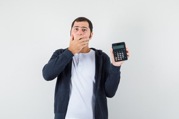 Young man holding calculator and covering mouth with hand in white t-shirt and zip-front black hoodie and looking optimistic , front view.