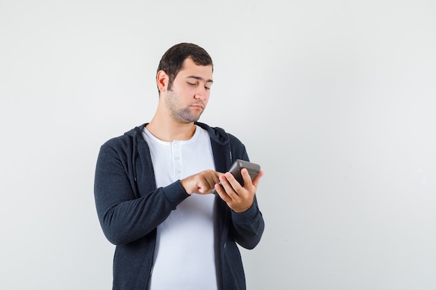 Young man holding calculator and doing some operations on it in white t-shirt and zip-front black hoodie and looking focused , front view.