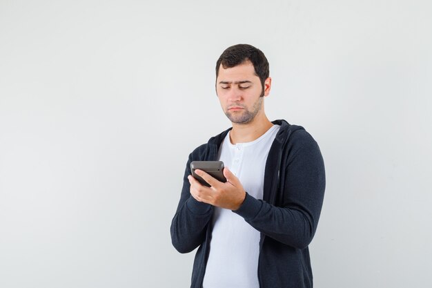 Young man holding calculator and doing some operations on it in white t-shirt and zip-front black hoodie and looking focused , front view.