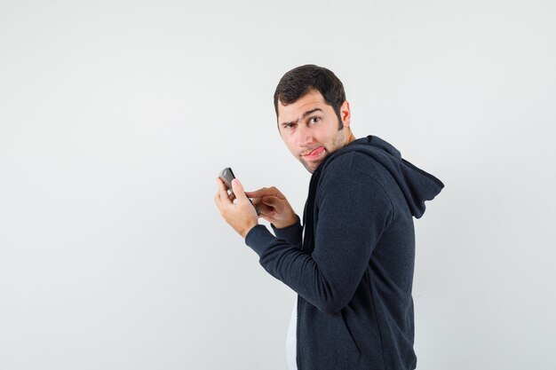 Young man holding calculator and doing some operations on it in white t-shirt and zip-front black hoodie and looking serious , front view.