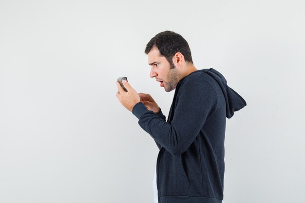 Young man holding calculator and doing some operations on it in white t-shirt and zip-front black hoodie and looking surprised , front view.