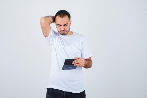 Young man holding calculator and holding hand behind head in white t-shirt and black pants and looking serious