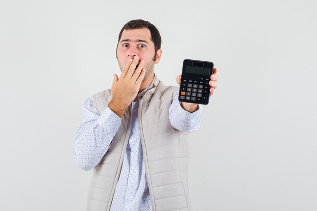 Young man holding calculator in one hand while covering mouth with hand in beige jacket and cap and looking surprised. front view.