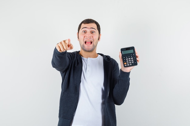 Young man holding calculator and pointing index finger at camera in white t-shirt and zip-front black hoodie and looking optimistic , front view.