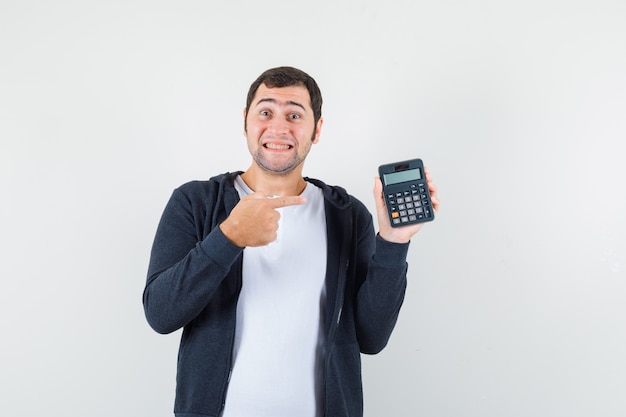 Young man holding calculator and pointing to it in white t-shirt and zip-front black hoodie and looking optimistic , front view.