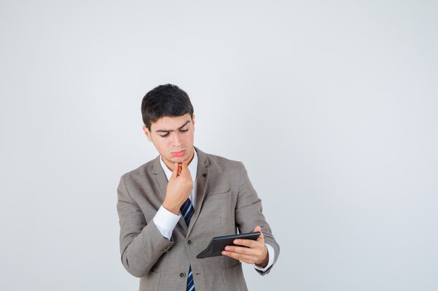 Young man holding calculator, putting hand on chin in formal suit