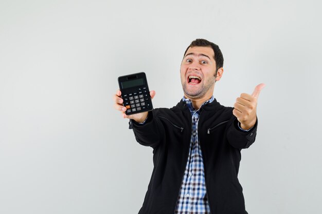 Молодой человек держит калькулятор, показывает палец вверх в рубашке, куртке и выглядит счастливым.