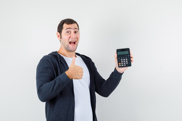 Young man holding calculator and showing thumb up in white t-shirt and zip-front black hoodie and looking optimistic , front view.