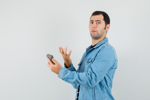 Young man holding calculator in t-shirt, jacket and looking puzzled .