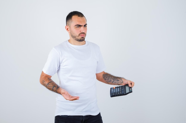 Young man holding calculator and thinking about something in white t-shirt and black pants and looking pensive
