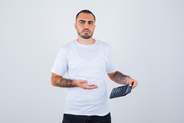 Молодой человек держит калькулятор и думает о чем-то в белой футболке и черных штанах и выглядит задумчивым