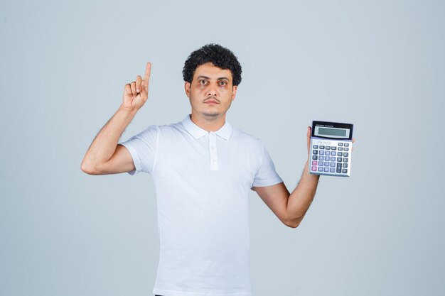 Молодой человек держит калькулятор, указывая вверх в белой футболке и выглядит серьезным. передний план.