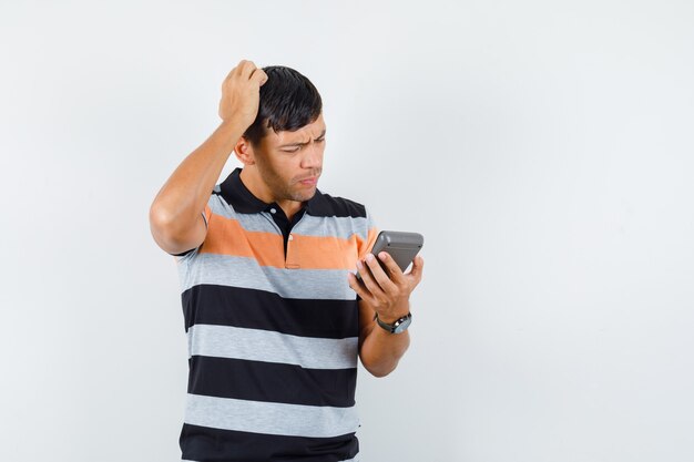 Young man holding calculator while scratching head in t-shirt and looking pensive