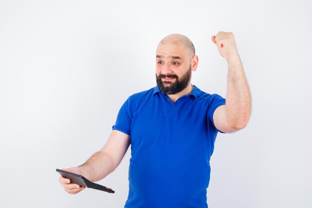 Young man holding calculator while showing success gesture in blue shirt and looking glad , front view.