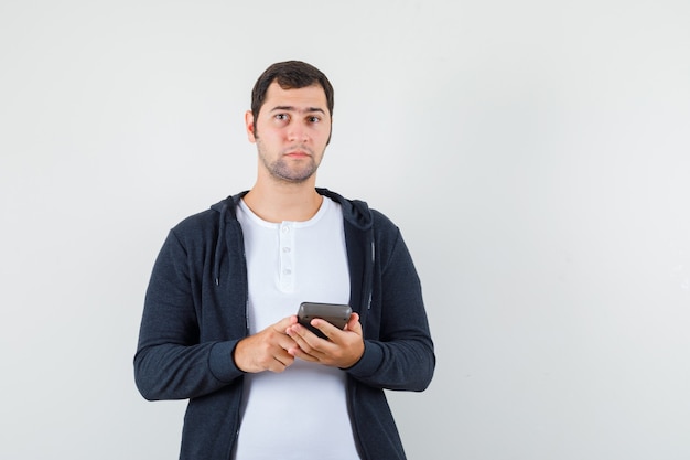 Young man holding calculator in white t-shirt and zip-front black hoodie and looking serious , front view.