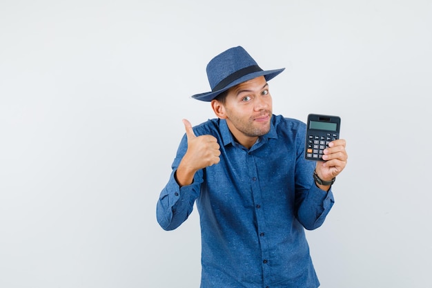 Молодой человек держит калькулятор с большим пальцем руки вверх в голубой рубашке, шляпе и смотрит оптимистично, вид спереди.