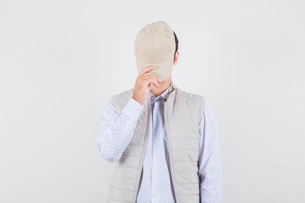 Young man holding cap on his face in shirt,sleeveless jacket and looking secret. front view.