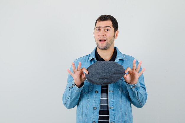 Young man holding cap in jacket,t-shirt and looking hesitant. 