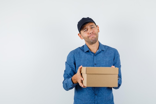 Young man holding cardboard box in blue shirt, cap and looking desperate , front view.