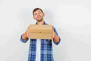 Free photo young man holding cardboard box in shirt and looking cheerful , front view.