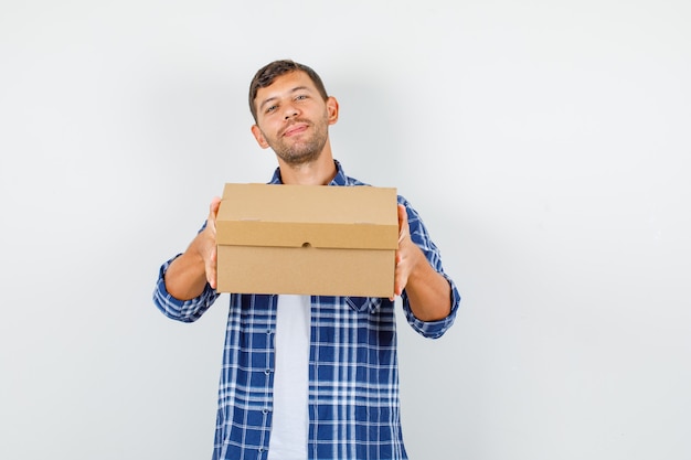 Young man holding cardboard box in shirt and looking cheerful , front view.