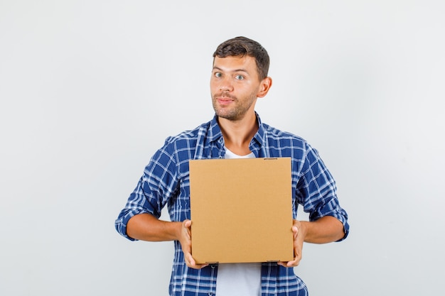 Young man holding cardboard box in shirt and looking positive , front view.