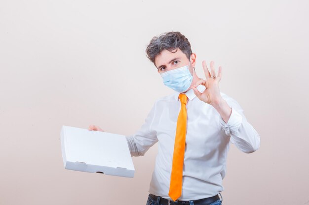 Young man holding cardboard box, showing ok sign in shirt, jeans, mask