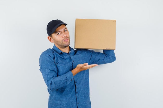 Young man holding cardboard box with spread palm in blue shirt, cap and looking glad. front view.