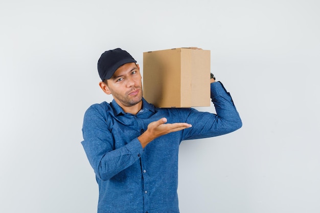 Young man holding cardboard box with spread palm in blue shirt, cap and looking glad. front view.