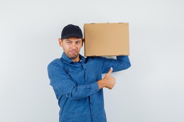 Young man holding cardboard box with thumb up in blue shirt, cap and looking glad. front view.