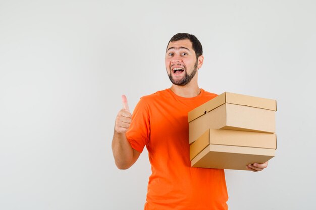 Молодой человек держит картонные коробки большим пальцем руки вверх в оранжевой футболке и смотрит оптимистично, вид спереди.