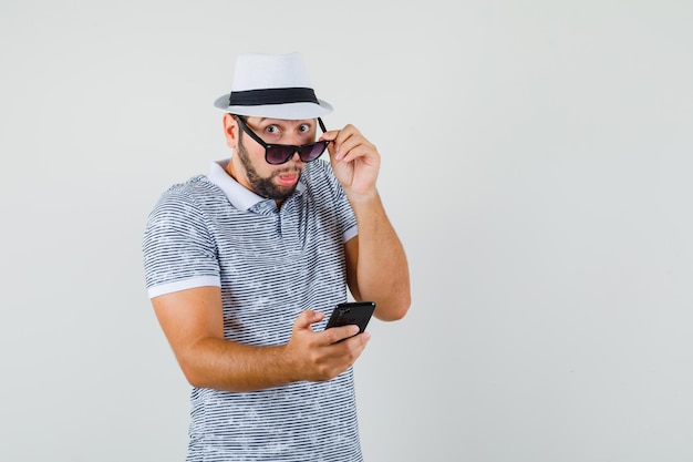 Young man holding cell phone, looking scrupulously over glasses in t-shirt, hat and looking puzzled , front view.