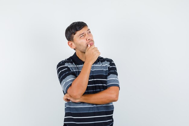 Young man holding chin while thinking in t-shirt and looking pensive. front view.