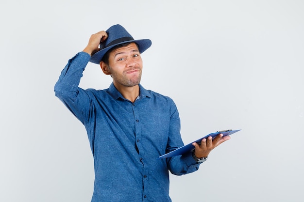 Young man holding clipboard in blue shirt, hat and looking hesitant , front view.