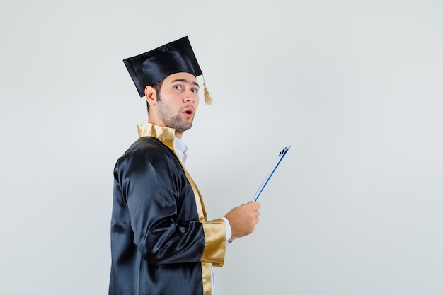 Young man holding clipboard in graduate uniform and looking worried.