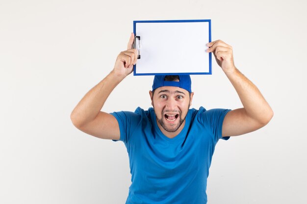 Young man holding clipboard above head in blue t-shirt and cap and looking successful