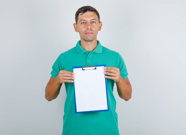 Young man holding clipboard and looking at camera in green t-shirt, front view.