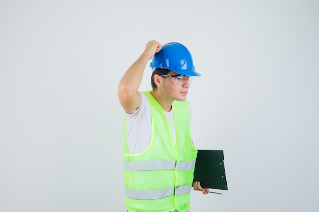 Young man holding clipboard and pen, putting hand on helmet in construction uniform and looking serious