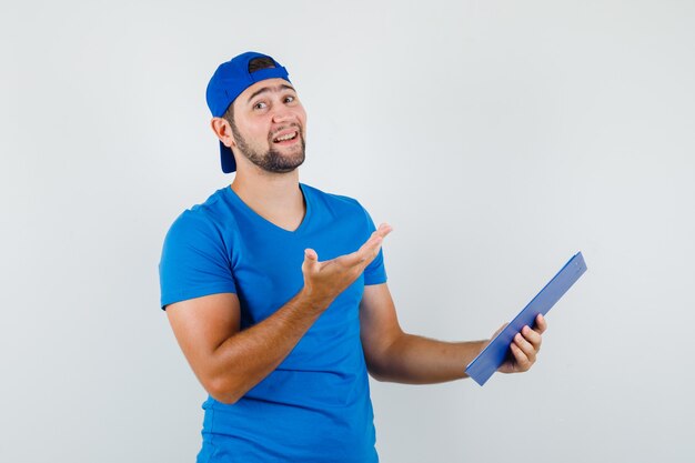 Young man holding clipboard and raising hand in blue t-shirt and cap and looking merry