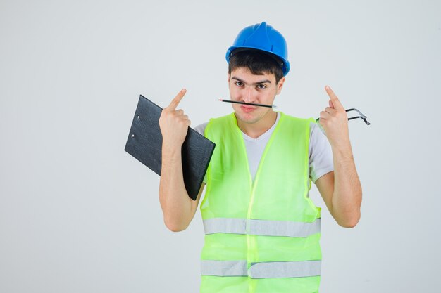 Young man holding clipboard and safety glasses in hands, holding pen with mouth