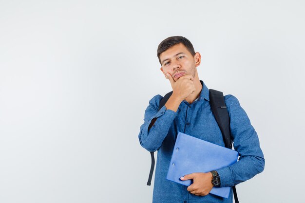 Young man holding clipboard with hand on chin in blue shirt and looking pensive. front view.