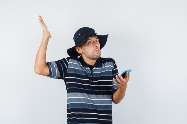 Young man holding clipboard with helpless gesture in t-shirt, hat and looking confused , front view.